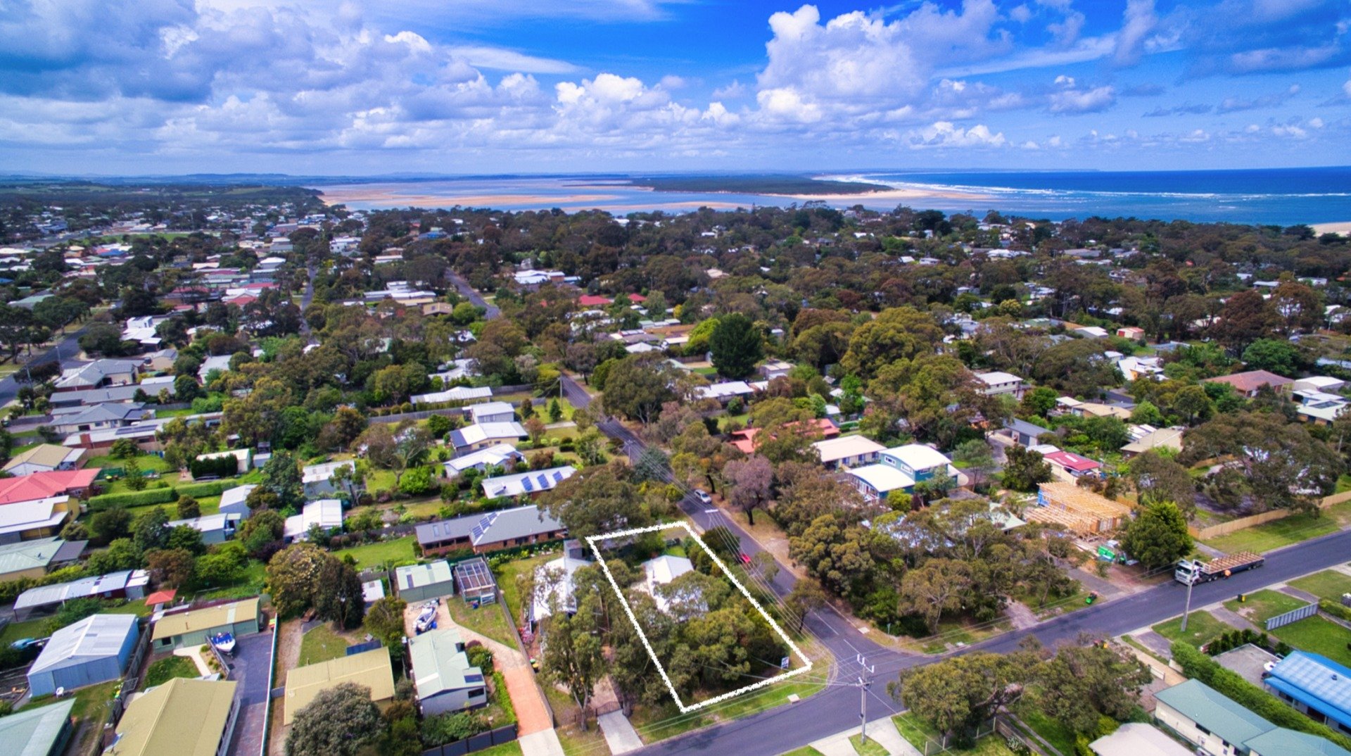 Aerial view of a suburban neighborhood with houses, trees, roads, and a distant coastline under a cloudy blue sky.