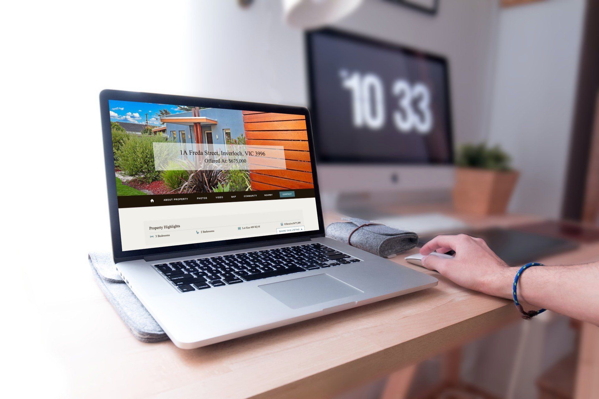 Laptop on desk showing a website, with a hand using a mouse beside a blurred monitor and keyboard.