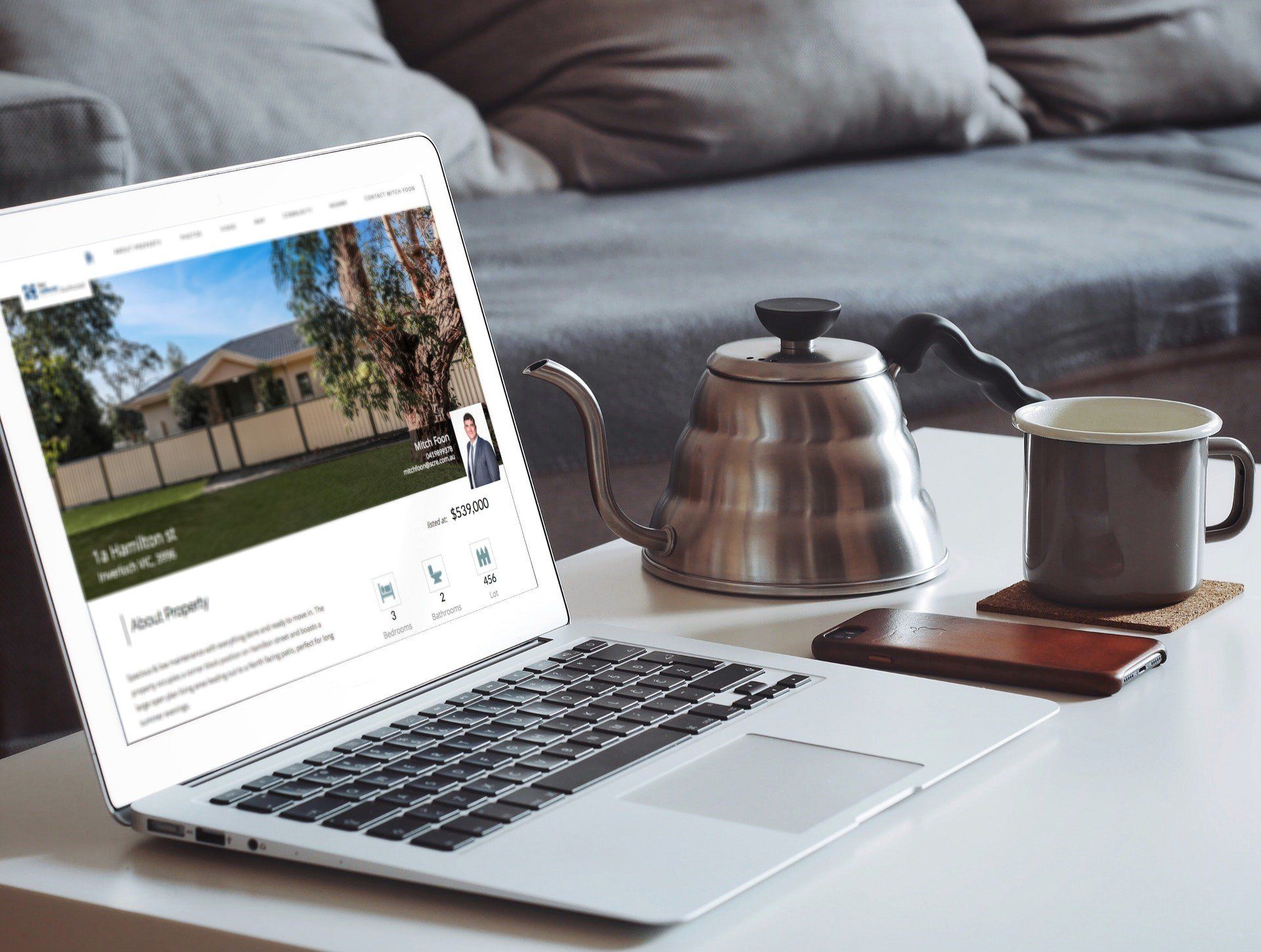 Laptop on a table beside a metal teapot and mug, showing a webpage with a house exterior.