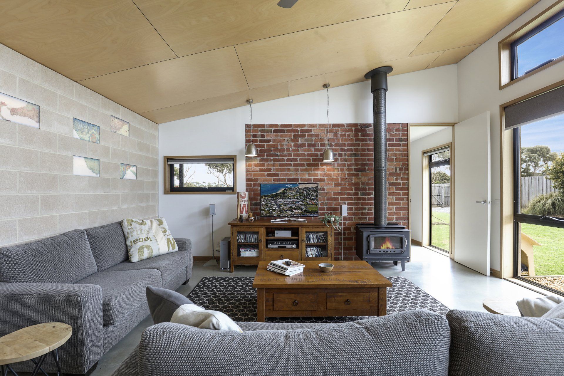 Modern living room with gray sofas, wooden coffee table, brick wall, and wood stove by large windows
