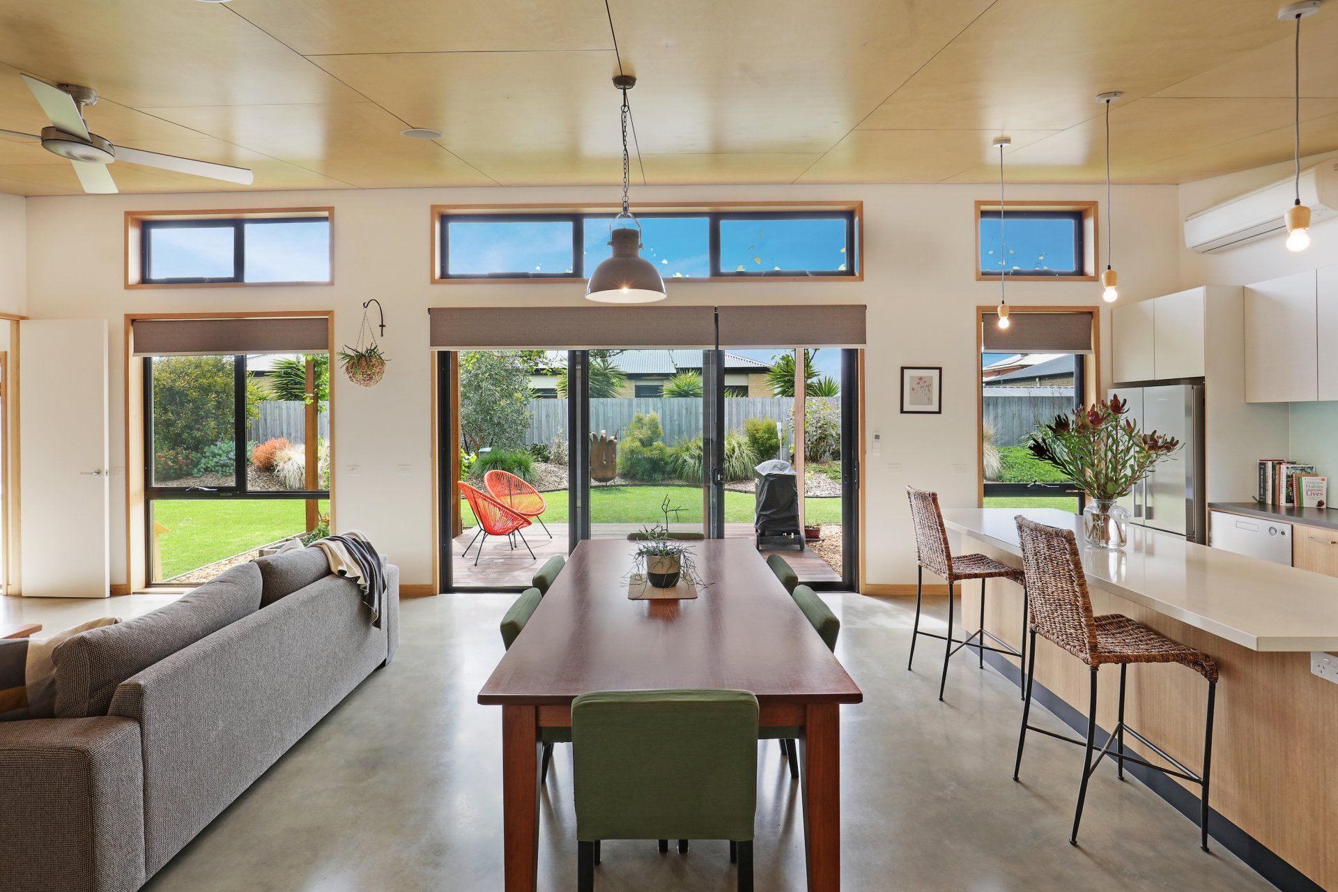 Modern open-plan kitchen and dining room with large windows, wooden table, bar stools, and garden views
