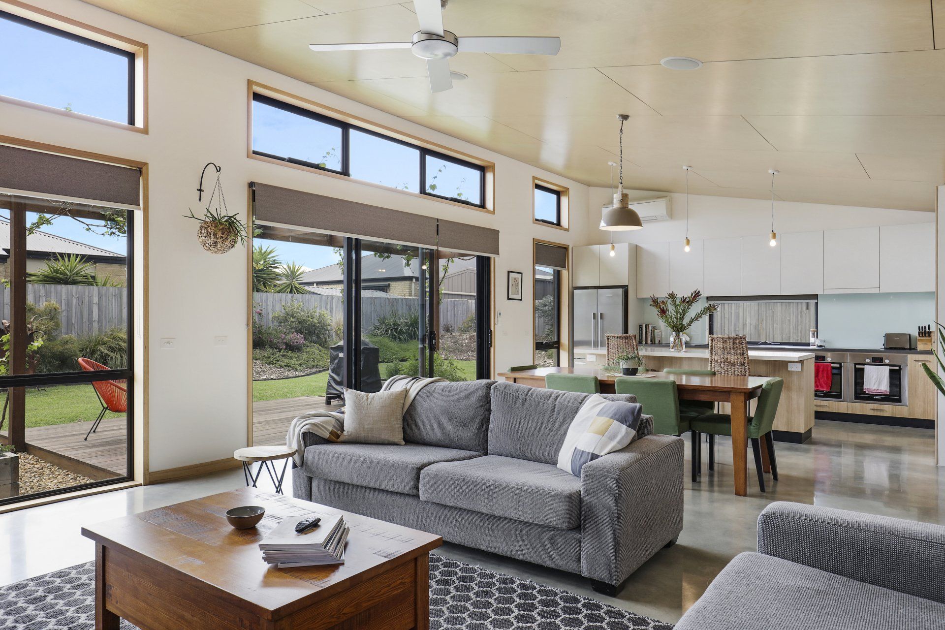 Bright modern living room with gray sofa, wooden table, open kitchen, and large windows overlooking outdoors