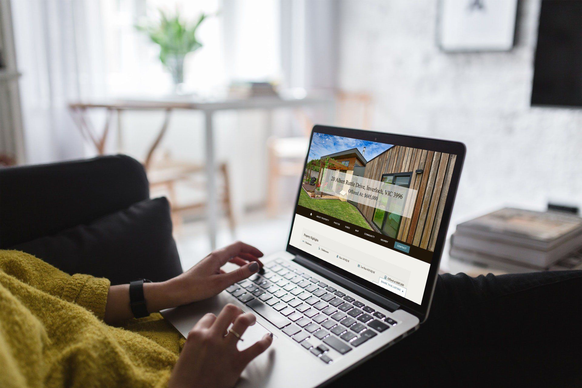 Person using a laptop on a couch in a bright living room, viewing a house listing.