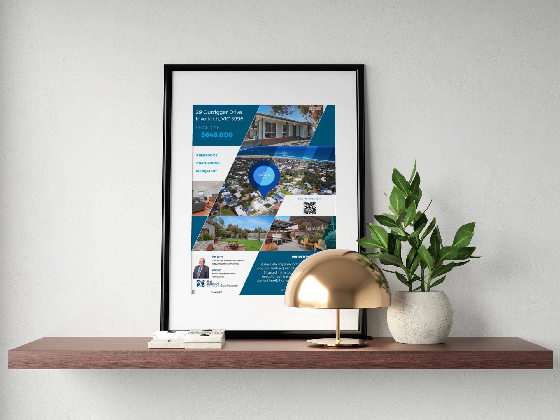 Framed travel poster on a white wall above a wooden shelf with a brass lamp and potted plant.