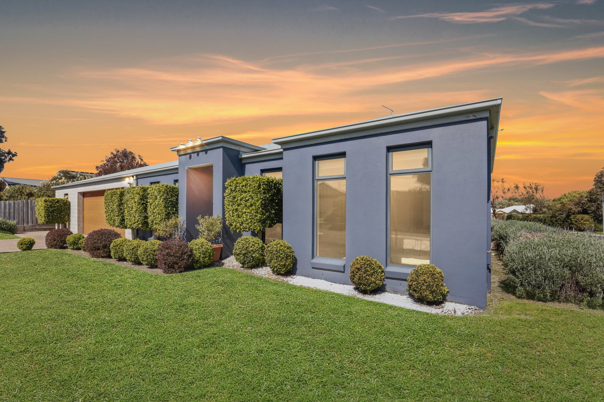 Modern single-story gray house with large windows, landscaped yard, and sunset sky