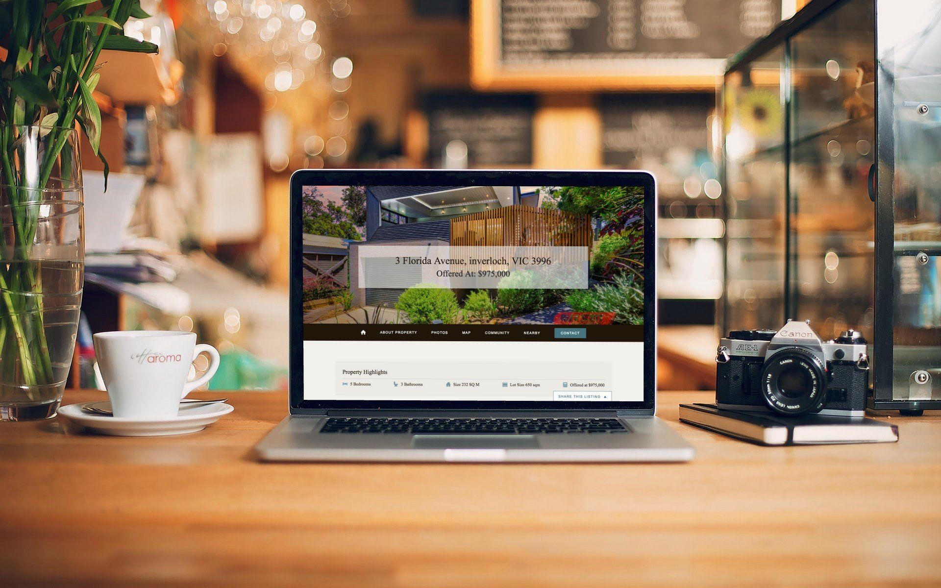Laptop on a wooden café table beside a camera and coffee cup, with a blurred warm-lit interior in the background