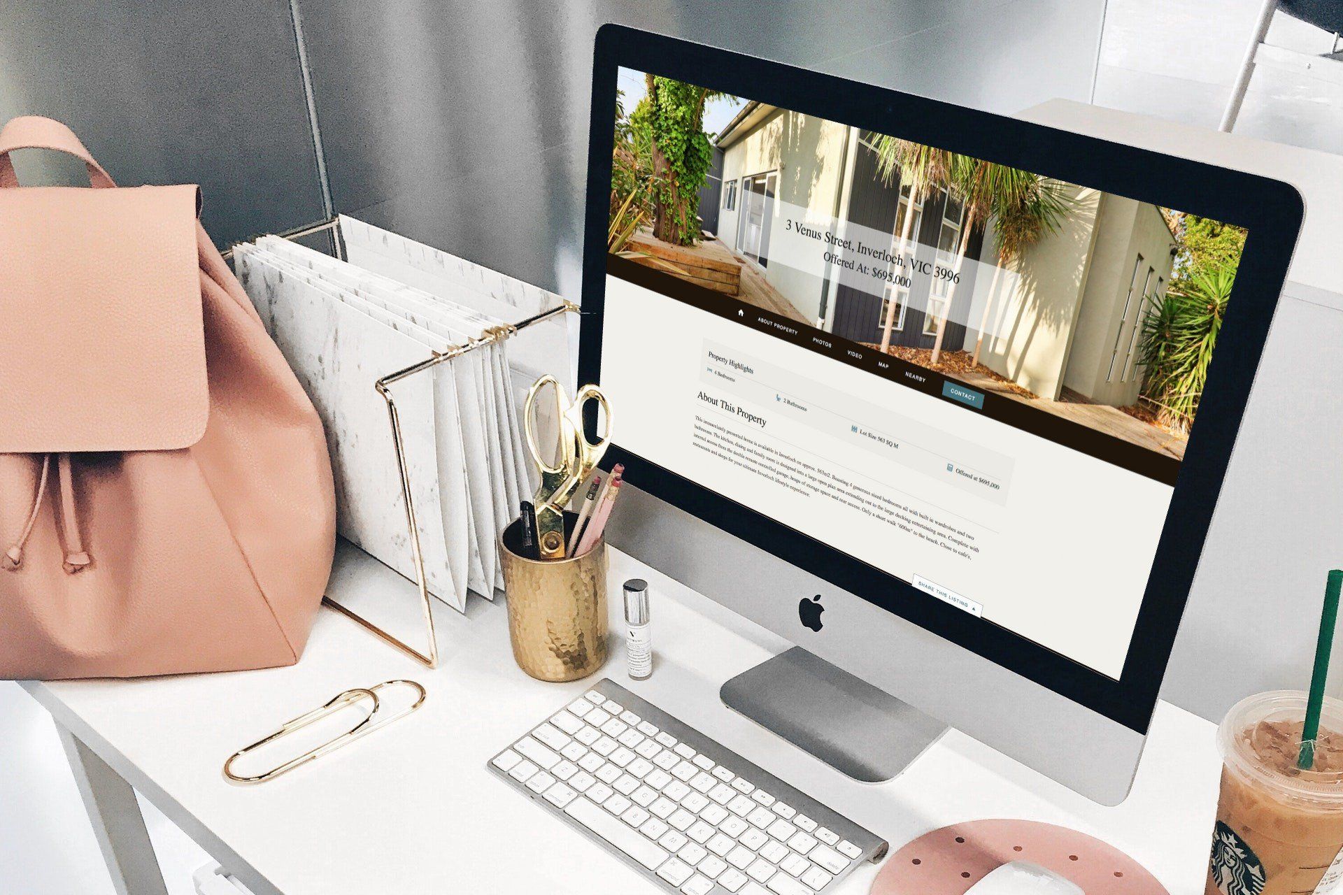 Minimal desk setup with iMac, pink bag, notebooks, and coffee on a white workspace