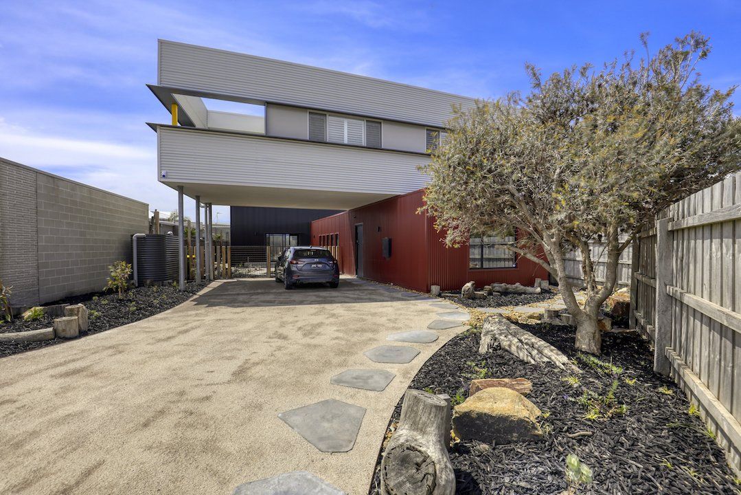 Modern house driveway with a car parked under an overhang and a red exterior wall, bordered by a tree and fence