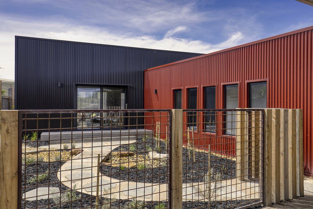 Modern modular building with black and rust-red corrugated walls and a fenced courtyard path