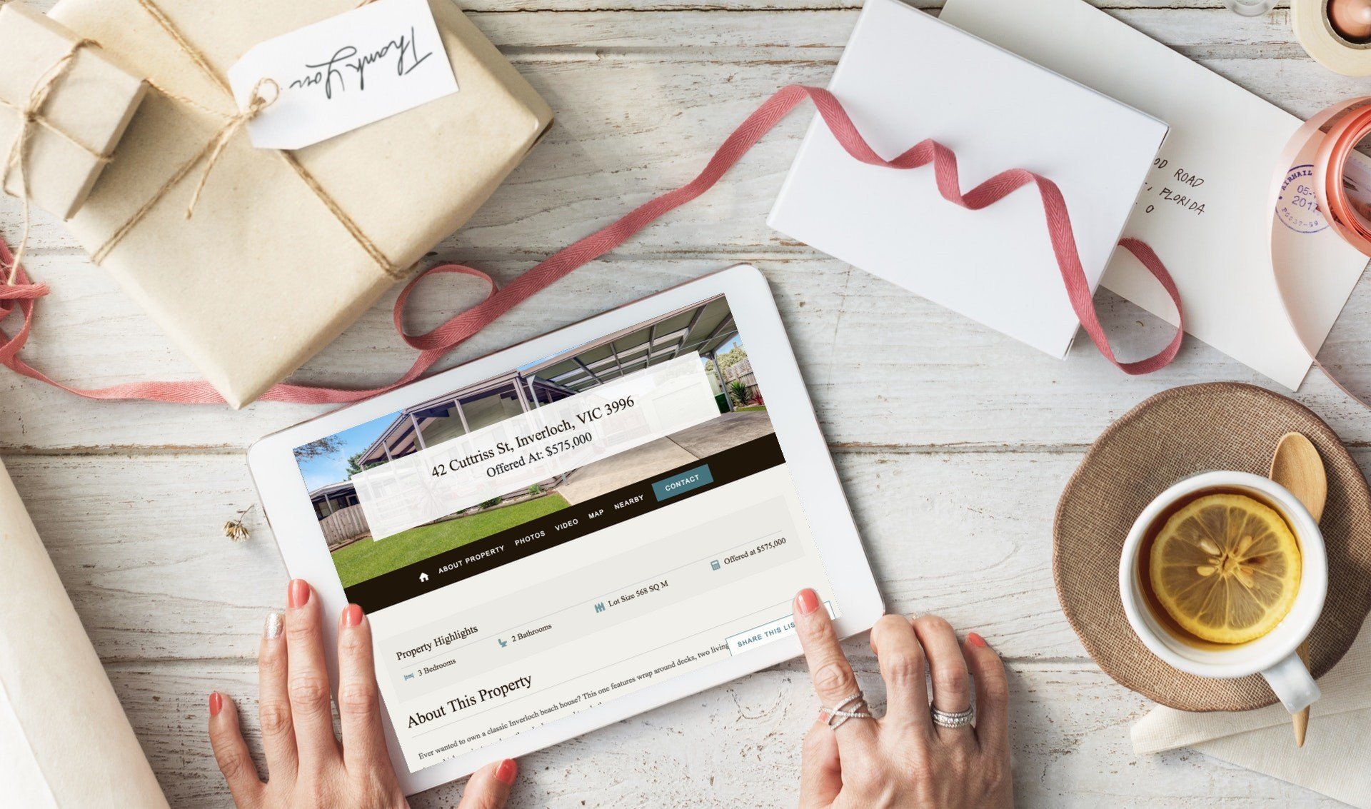 Hands holding a tablet beside wrapped gifts, a card, and a cup of tea on a white wooden table.