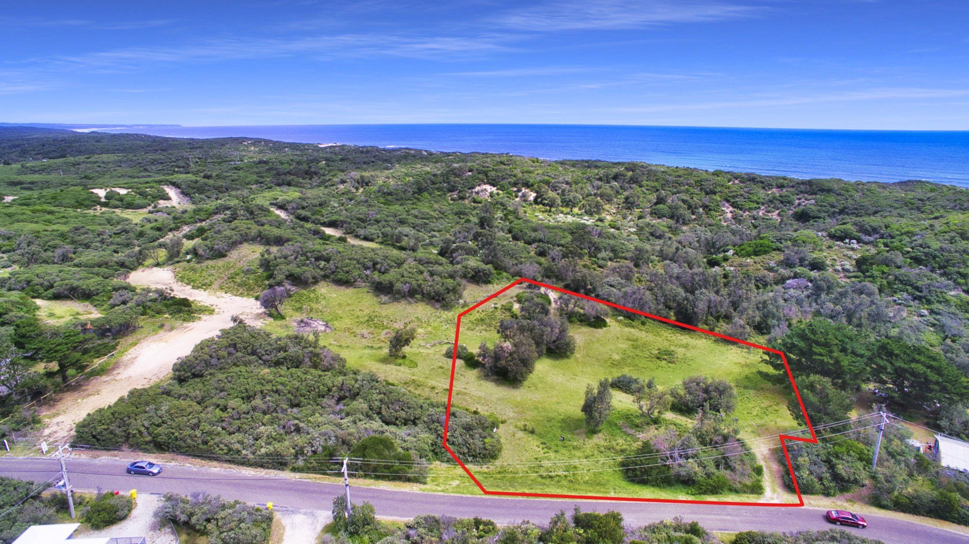Coastal landscape with a red-outlined grassy lot beside a road and dense green vegetation under a blue sky