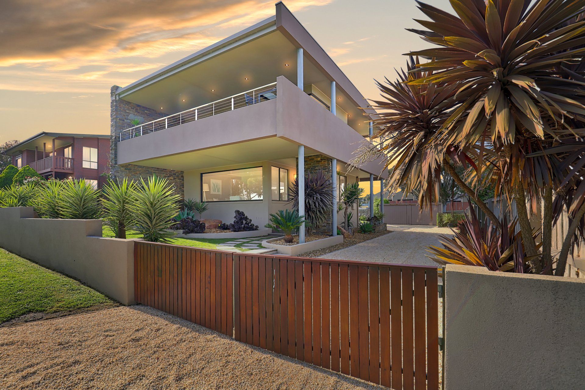 Modern two-story house with balcony, front garden, and wooden fence at sunset