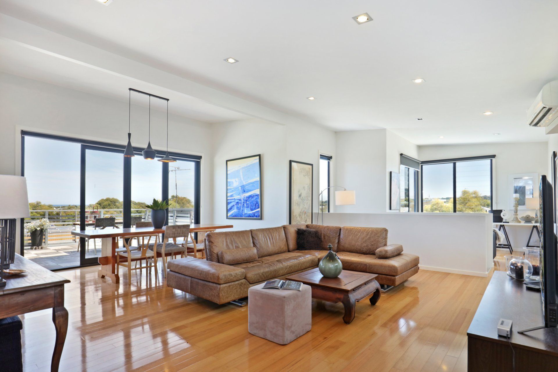 Bright modern living room with brown sectional sofa, wooden floors, and large windows overlooking a dining area