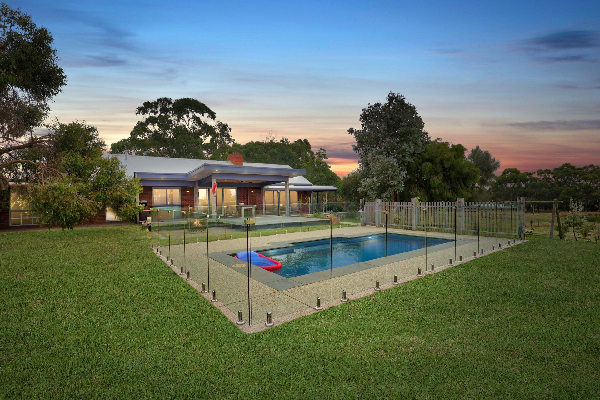 Backyard pool at dusk with glass fence, green lawn, and a house glowing under a pink sunset sky