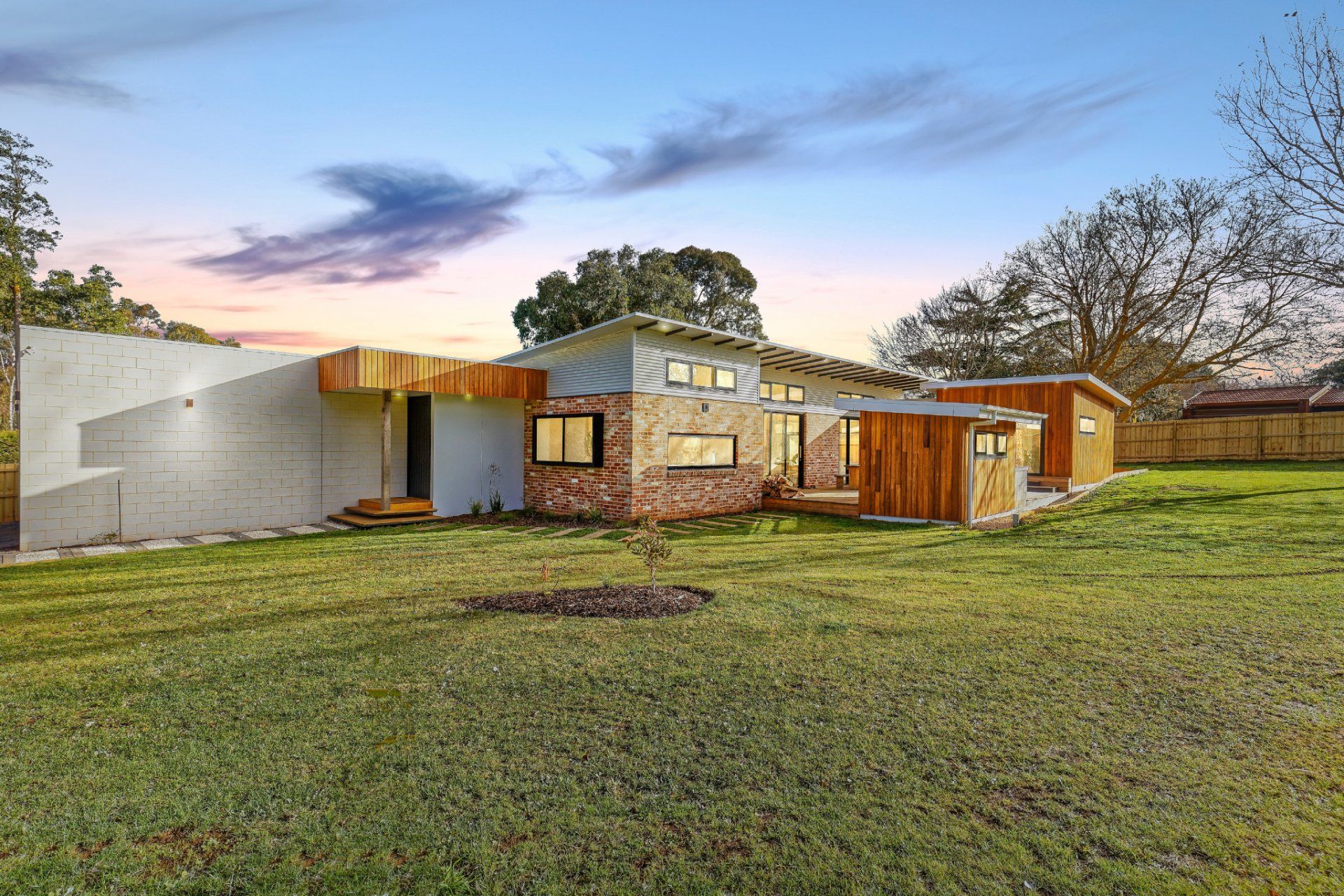 Modern single-story house with stone and wood exterior, a large grassy yard, and sunset sky.