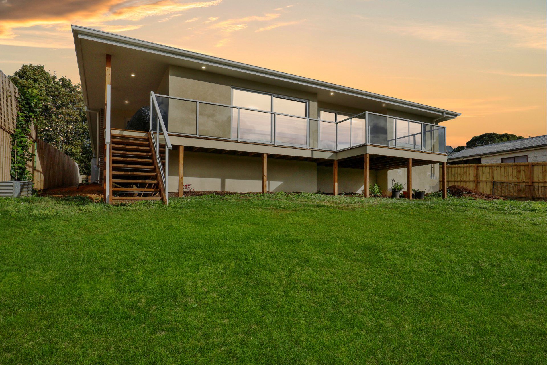Modern elevated house with a covered deck, stairs, and a large green lawn at sunset