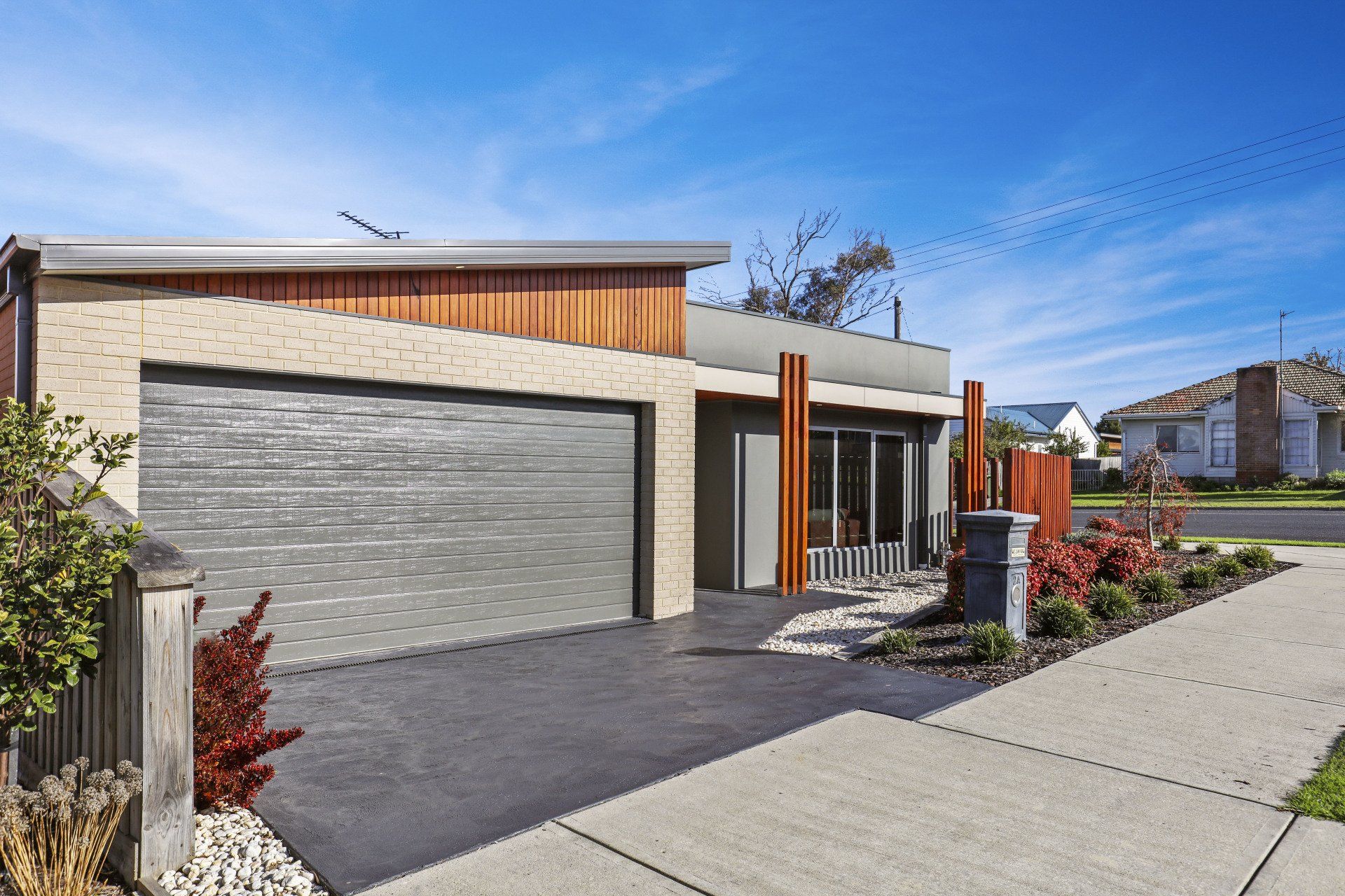 Modern single-story house with a gray garage door, stone facade, and front sidewalk under a blue sky