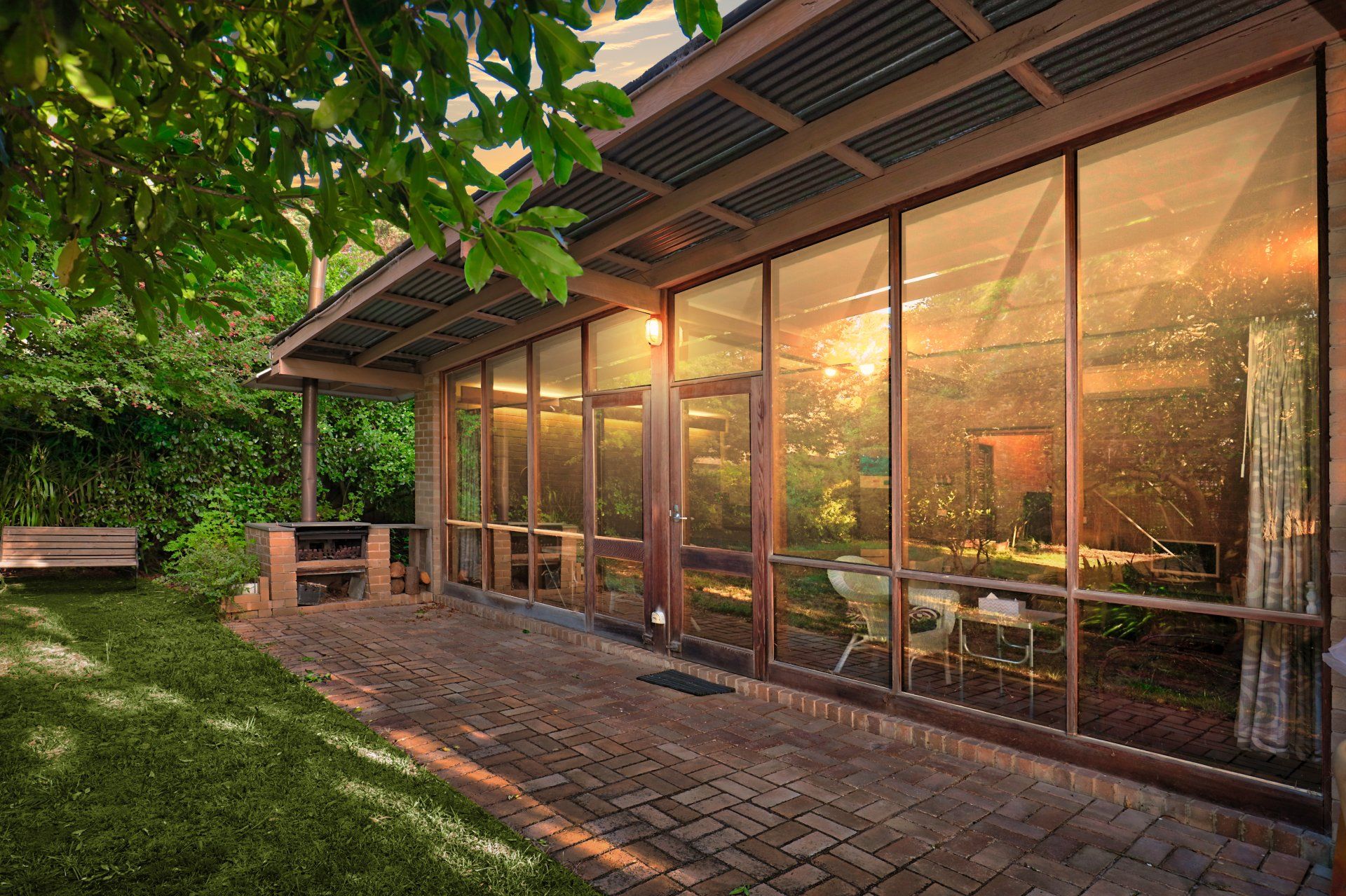 Glass-walled patio/café at sunset with warm lights, brick path, and surrounding green trees