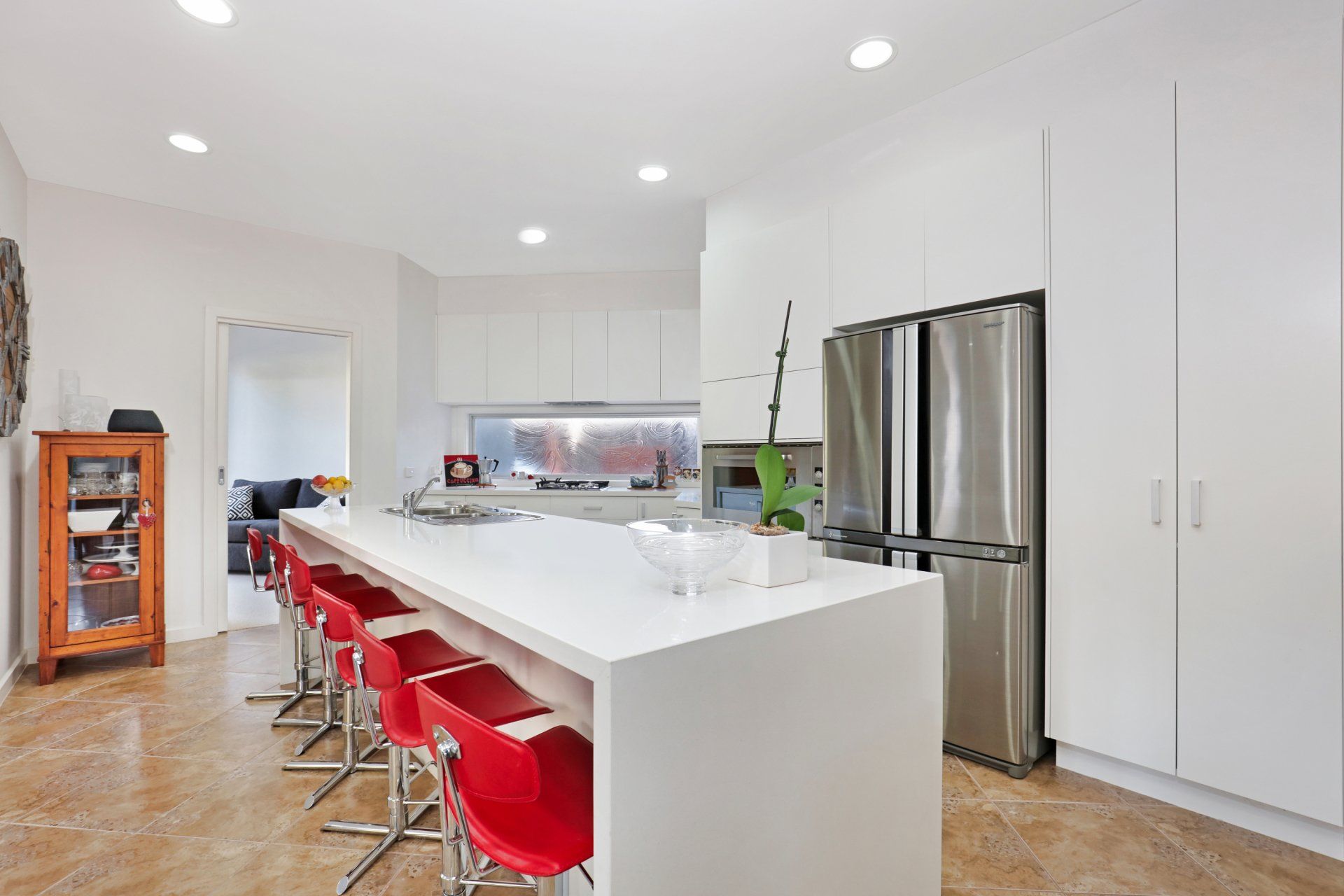 Modern white kitchen with large island, red stools, and stainless steel refrigerator