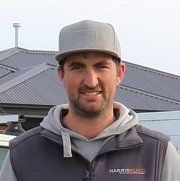 Man in a gray cap and hoodie standing outdoors in front of a roof, looking at the camera.