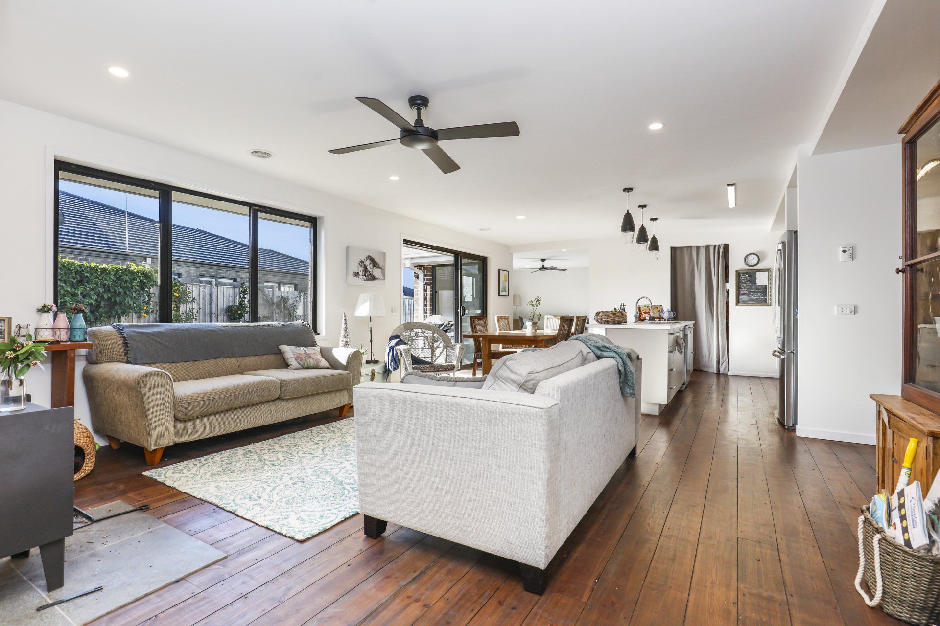 Bright open living room with white sofa, brown couch, wood floors, ceiling fan, and kitchen beyond.
