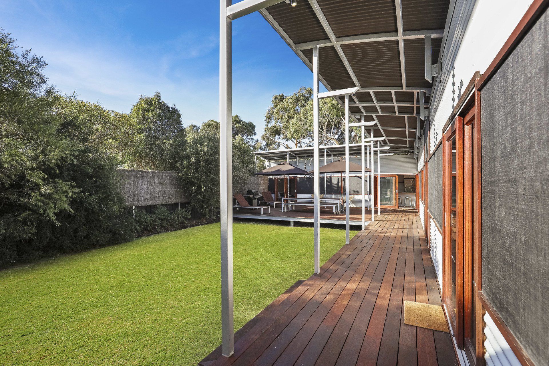Covered wooden deck beside a green lawn, with white posts and a corrugated roof along a brick wall.
