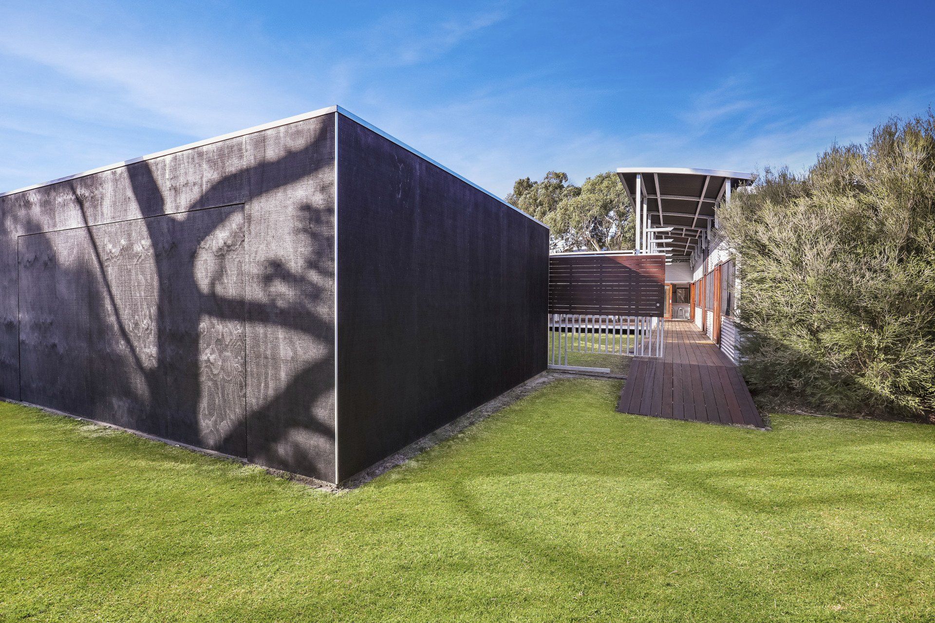 Modern building with a black textured wall, grassy lawn, and a wooden ramp under a blue sky.
