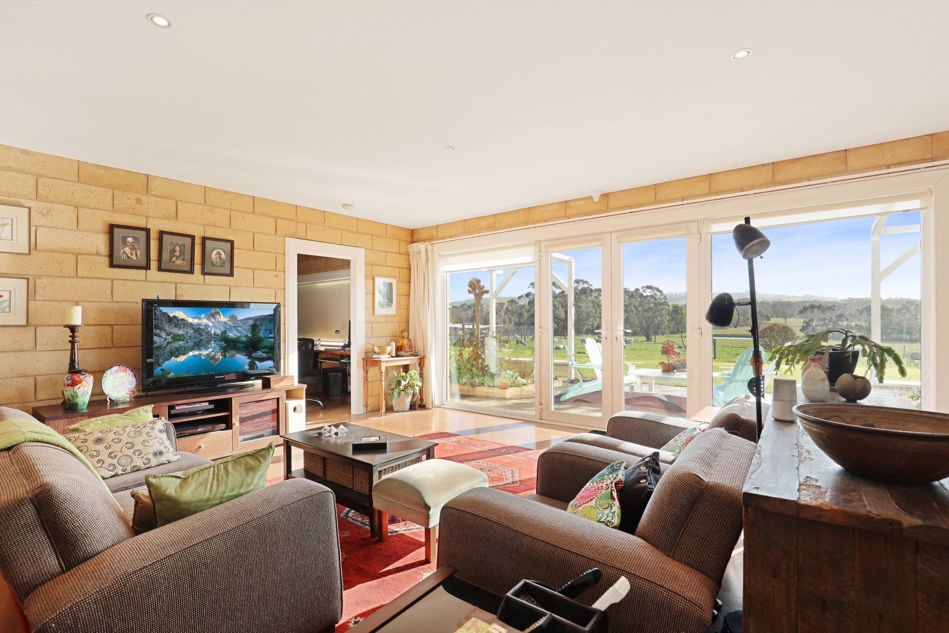 Bright living room with brown sofas, TV, red rug, and large windows overlooking a sunny landscape