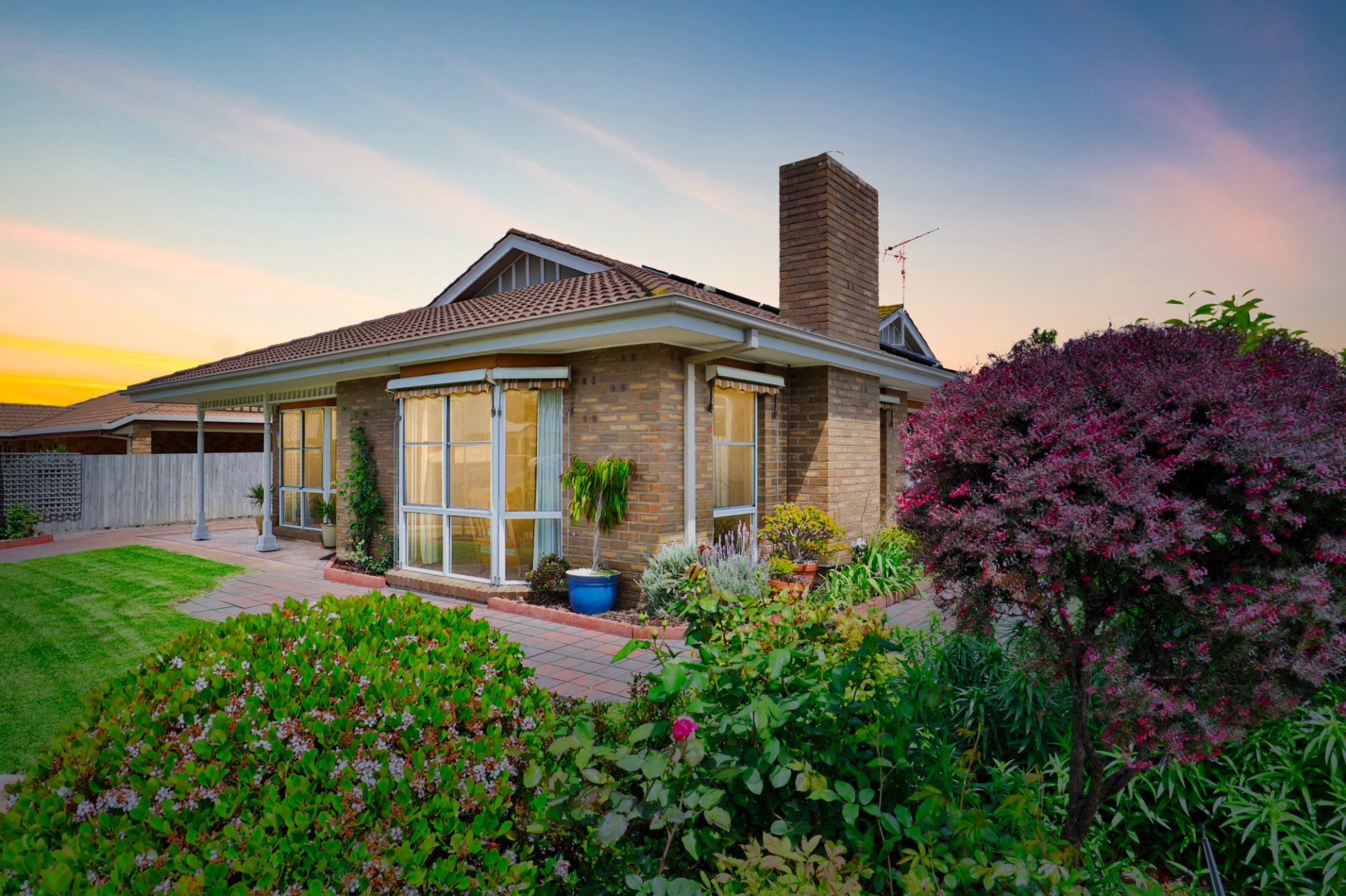 Brick house with a tiled roof, lit at sunset, surrounded by lush green and purple landscaping.