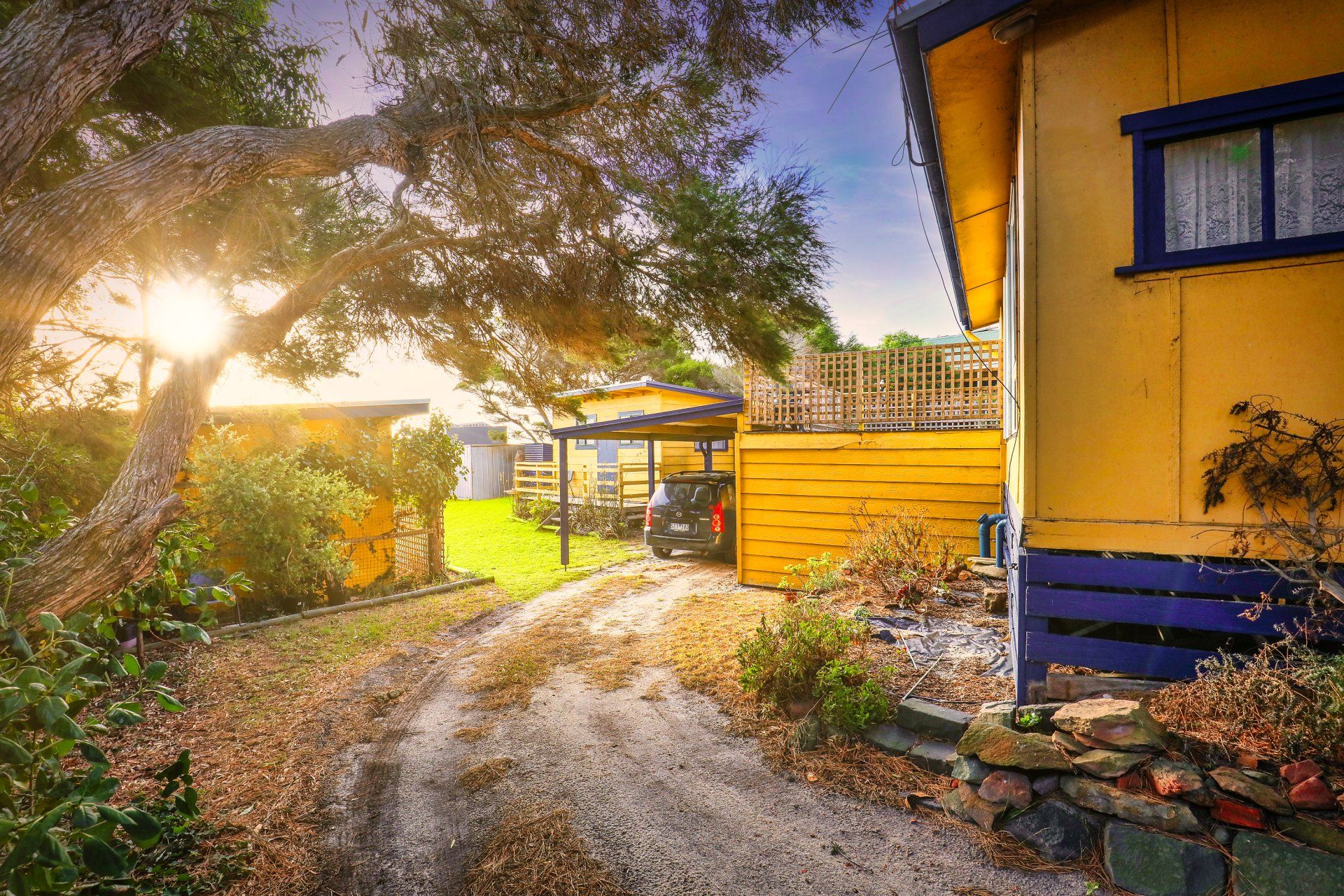 Sunlit gravel path beside a yellow house and trees at dusk