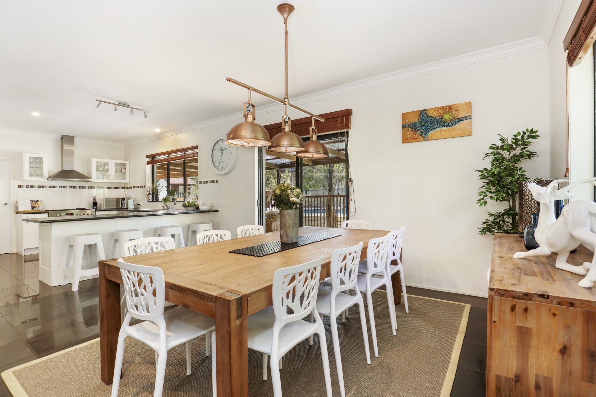 Bright dining room with wooden table, white chairs, and open kitchen with island in the background
