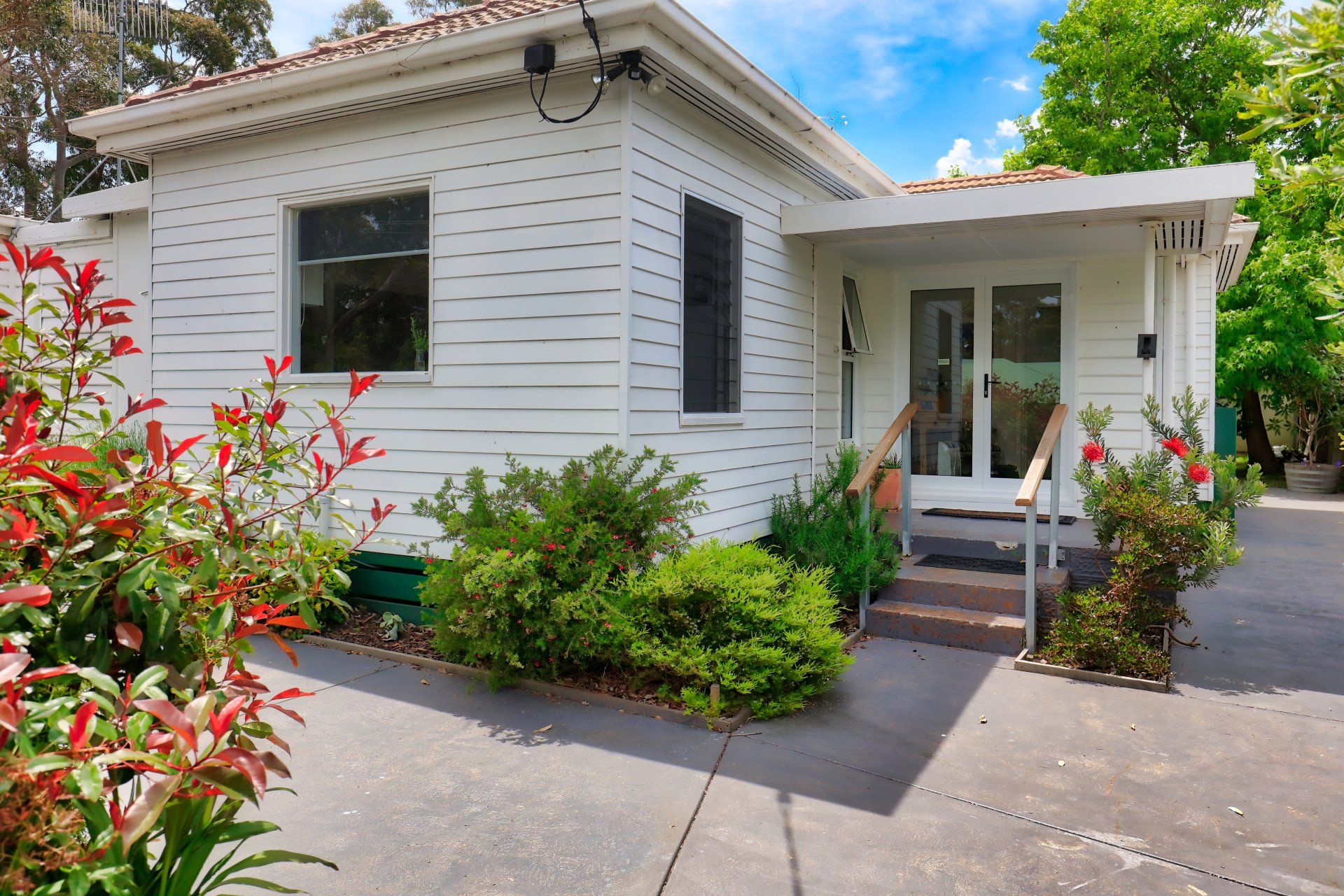 White single-story house with a front porch, garden shrubs, and a concrete driveway under a blue sky