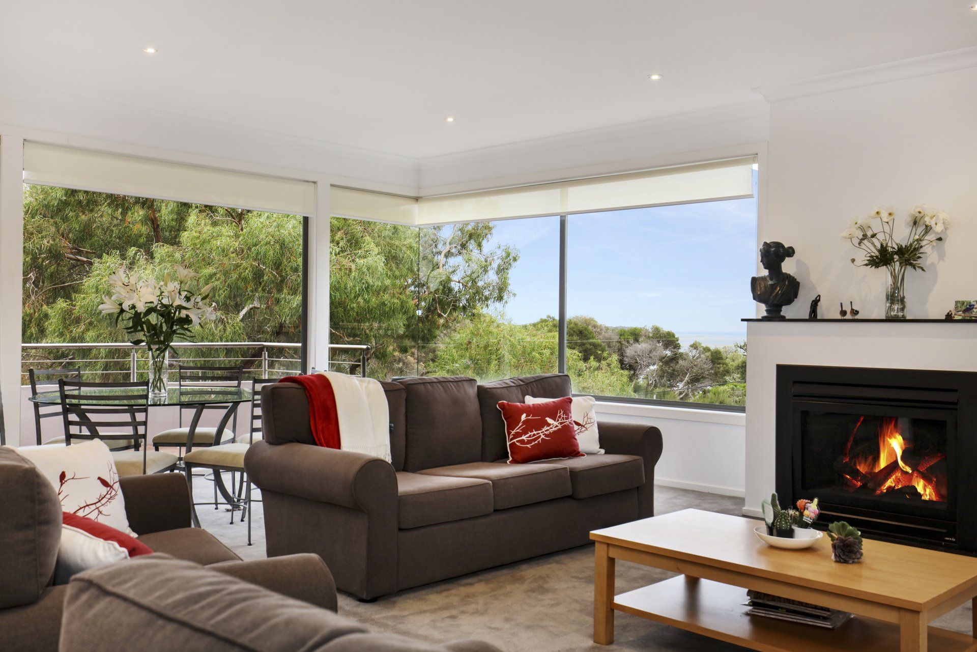 Bright living room with large windows, dark sofa, coffee table, and lit fireplace overlooking greenery