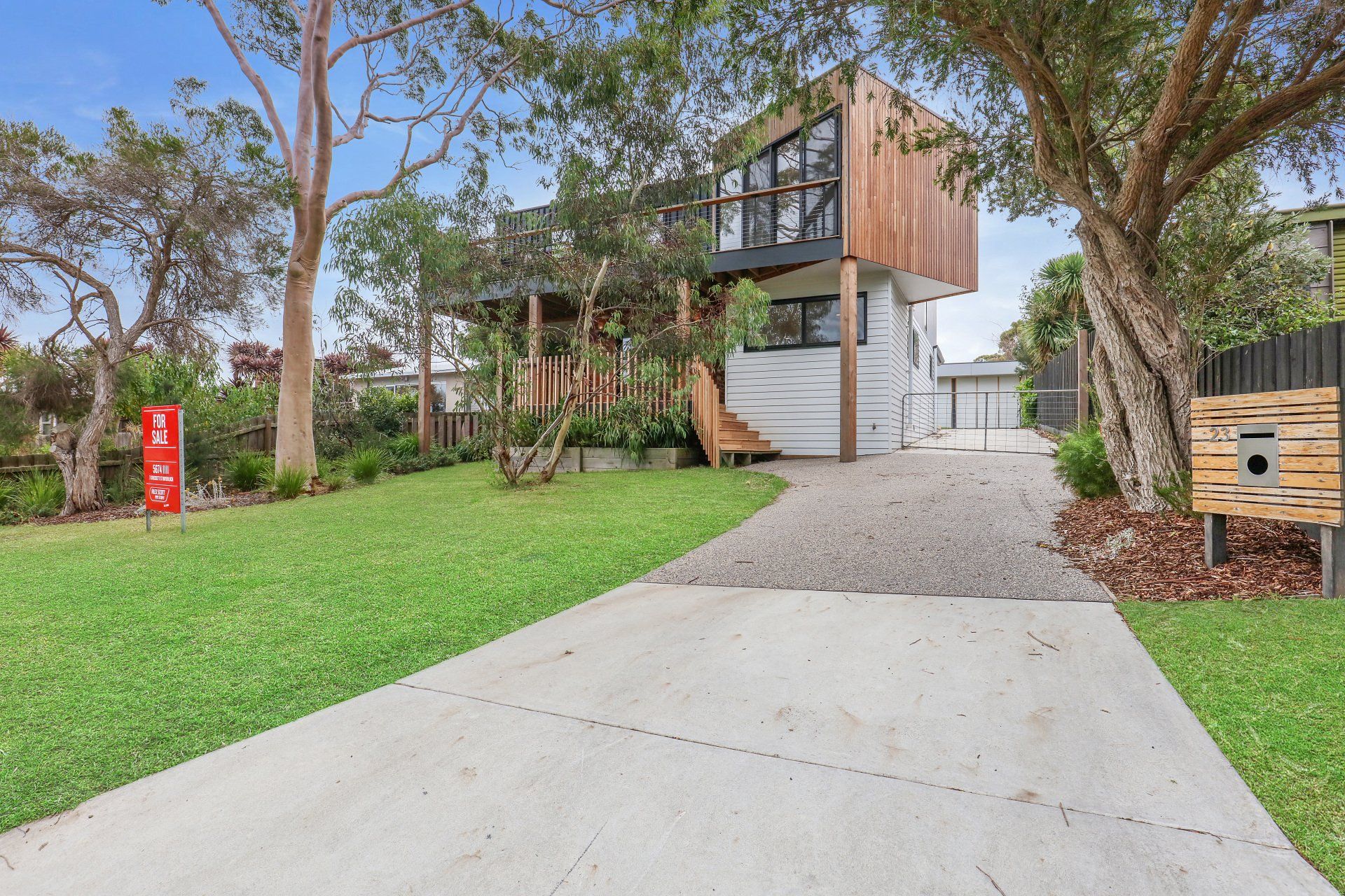Modern house with wooden balcony, gravel driveway, and green lawn among trees