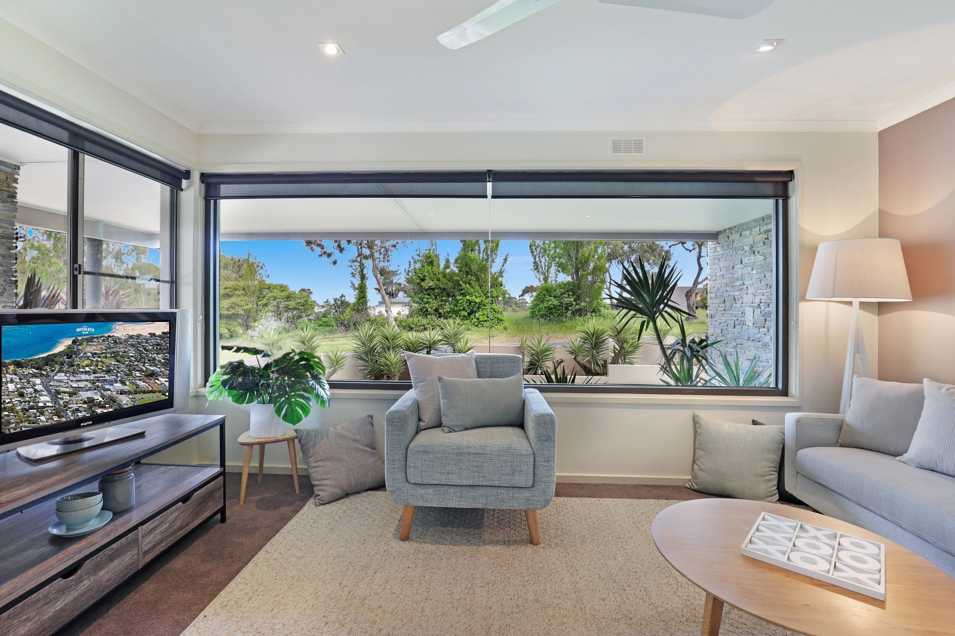 Bright living room with large windows, gray armchair, white sofa, and leafy outdoor view.