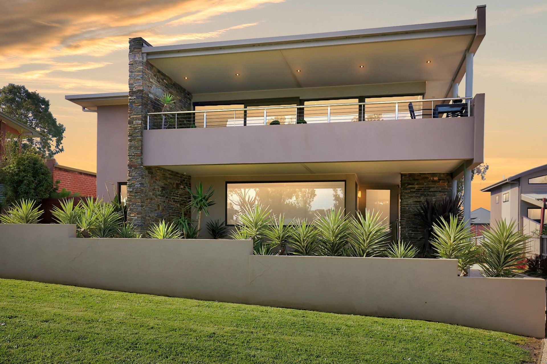 Modern two-story house with large windows, balcony, and a landscaped front yard at sunset