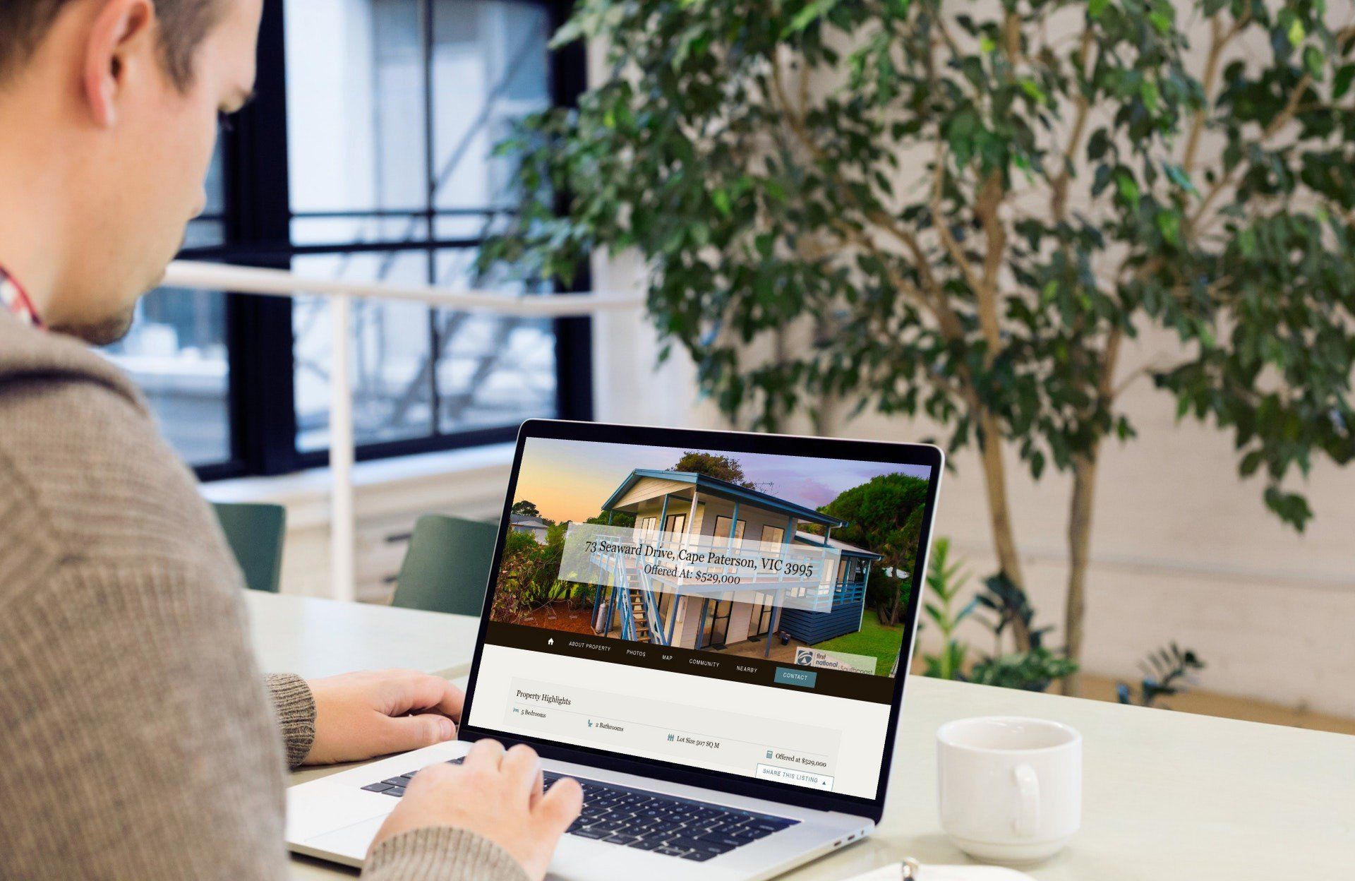 Person using a laptop at a table in a bright office with a large plant nearby