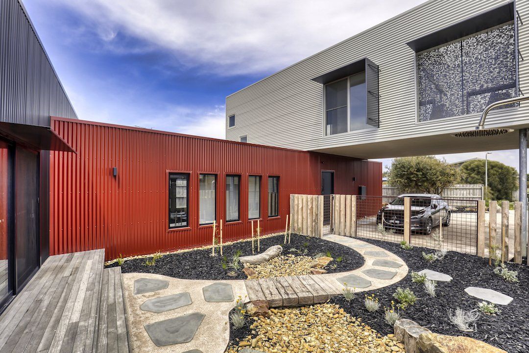 Modern courtyard with red and gray buildings, stone path, circular planter, and parked car under an overhang