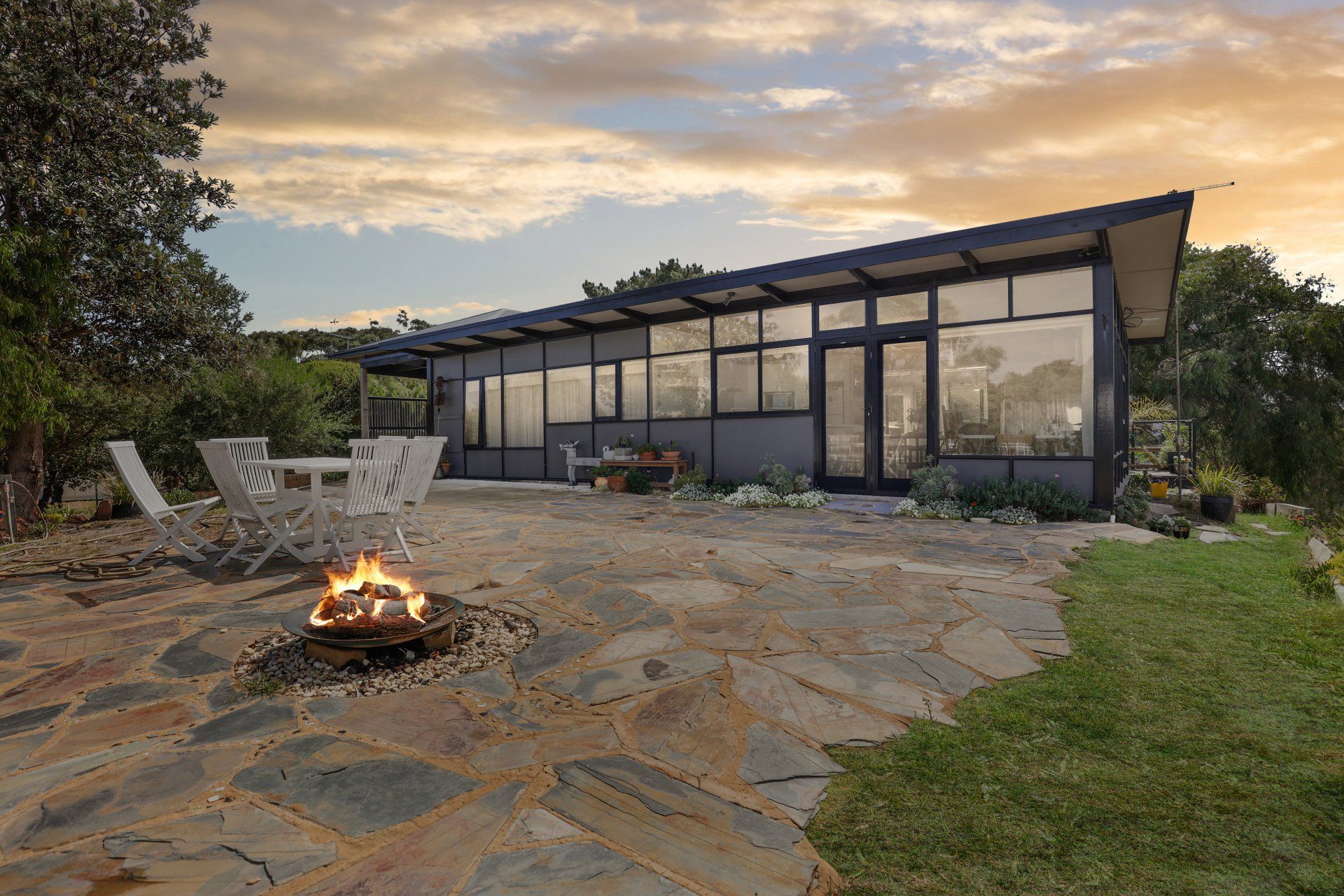 Modern glass house with stone patio, fire pit, and lawn at dusk