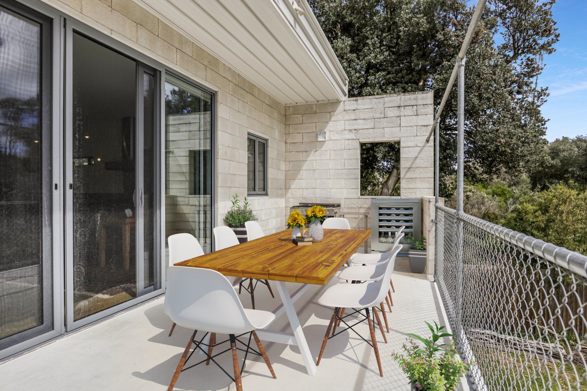 White patio with wooden table, white chairs, sliding glass doors, and a chain-link railing overlooking trees