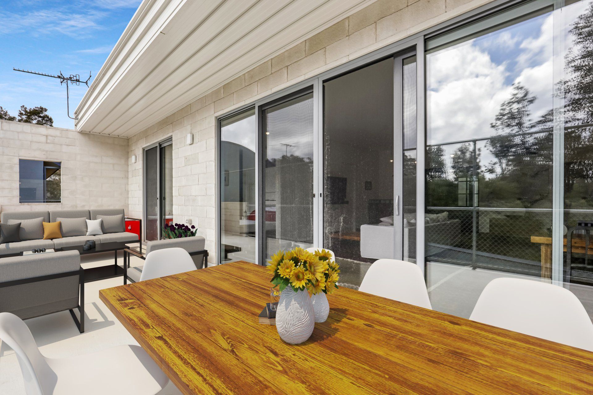 Modern patio with wooden table, white chairs, gray sofa, and sliding glass doors overlooking greenery