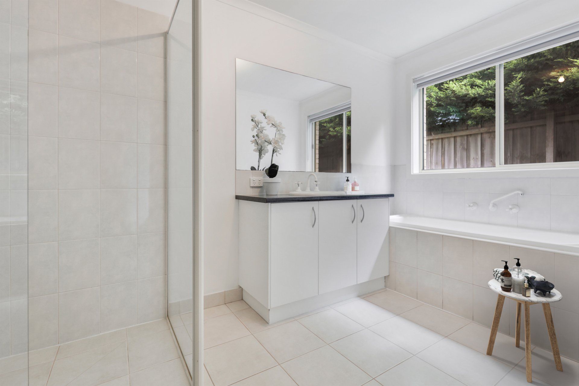 Bright modern bathroom with glass shower, white vanity, window, and beige tile accents