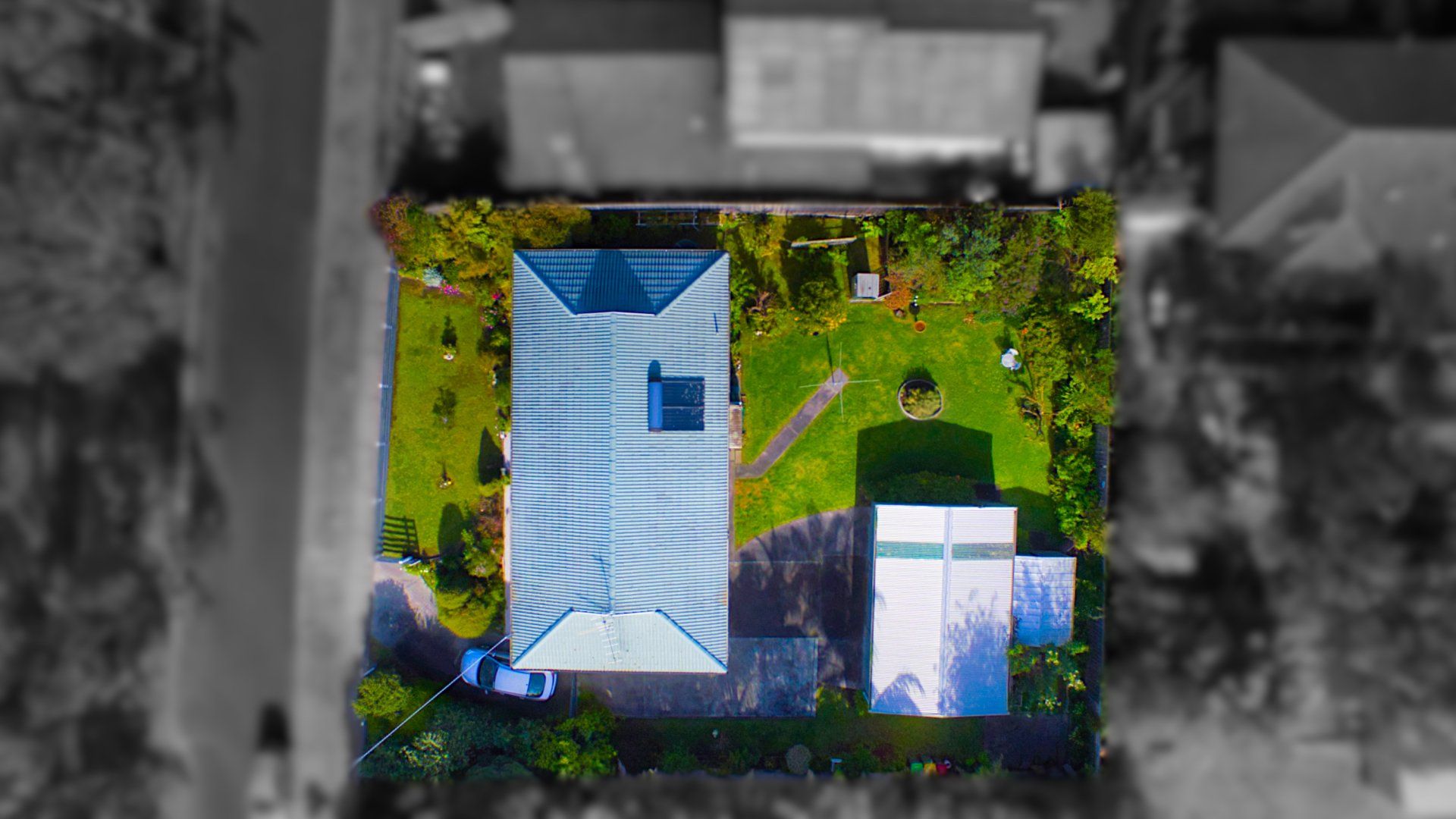 Aerial view of a blue-roofed house with a driveway, surrounded by trees and greenery.