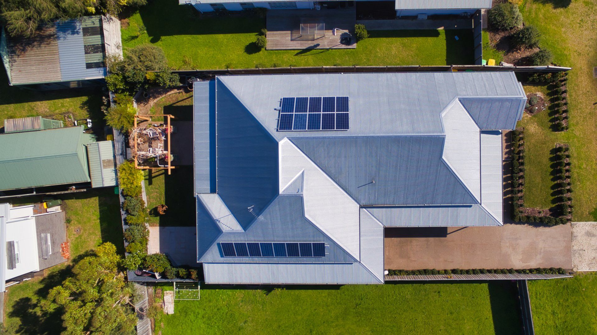 Aerial view of a house with a gray metal roof, solar panels, and a driveway surrounded by green lawns.