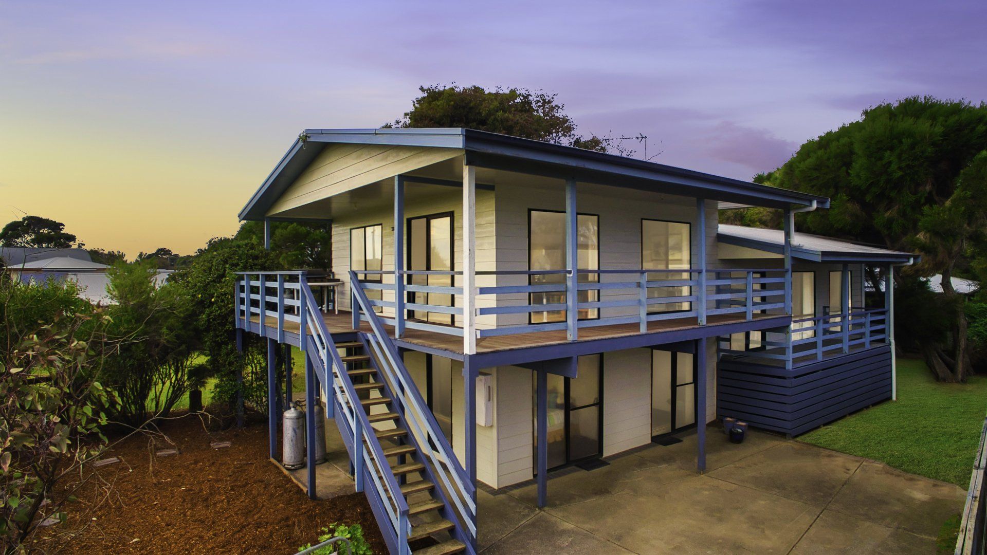 Elevated blue house with wraparound porch and stairs at dusk in a grassy yard