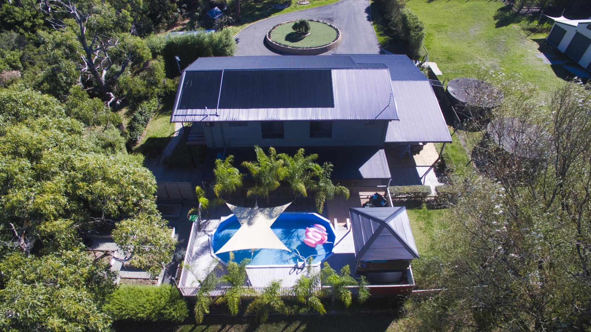 Aerial view of a house with solar panels, a pool, and patio umbrellas surrounded by trees