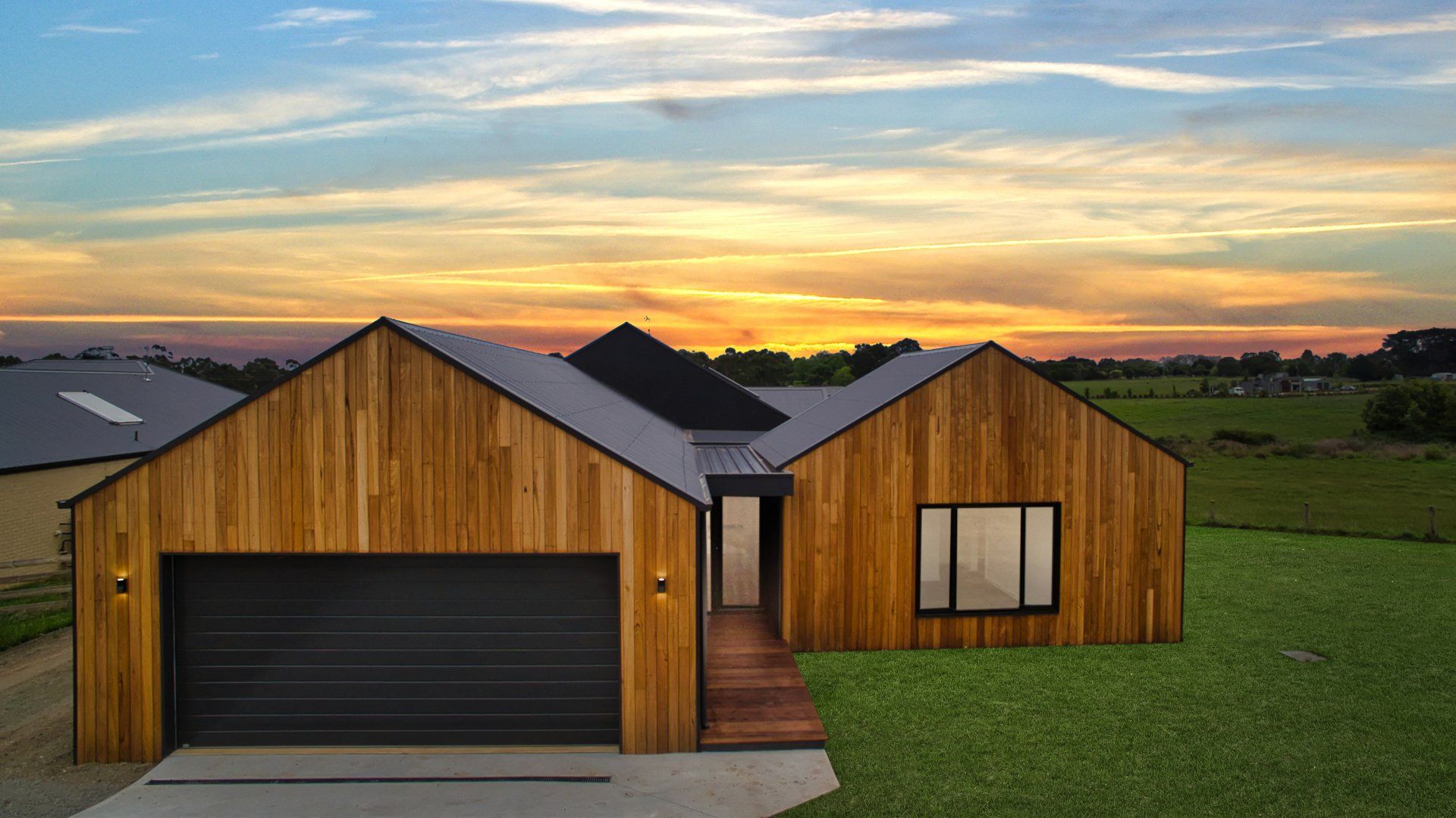 Modern wooden house with a garage at sunset, set in a grassy field under a colorful sky