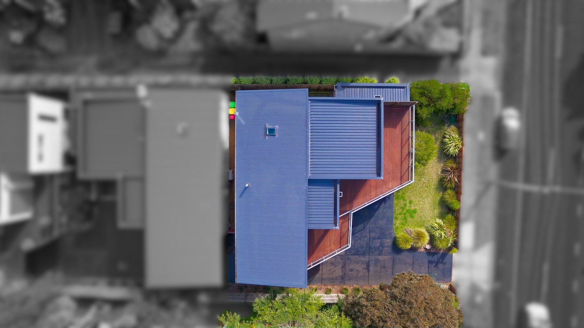 Aerial view of a blue-roofed house with a red deck, surrounded by trees and neighboring buildings.