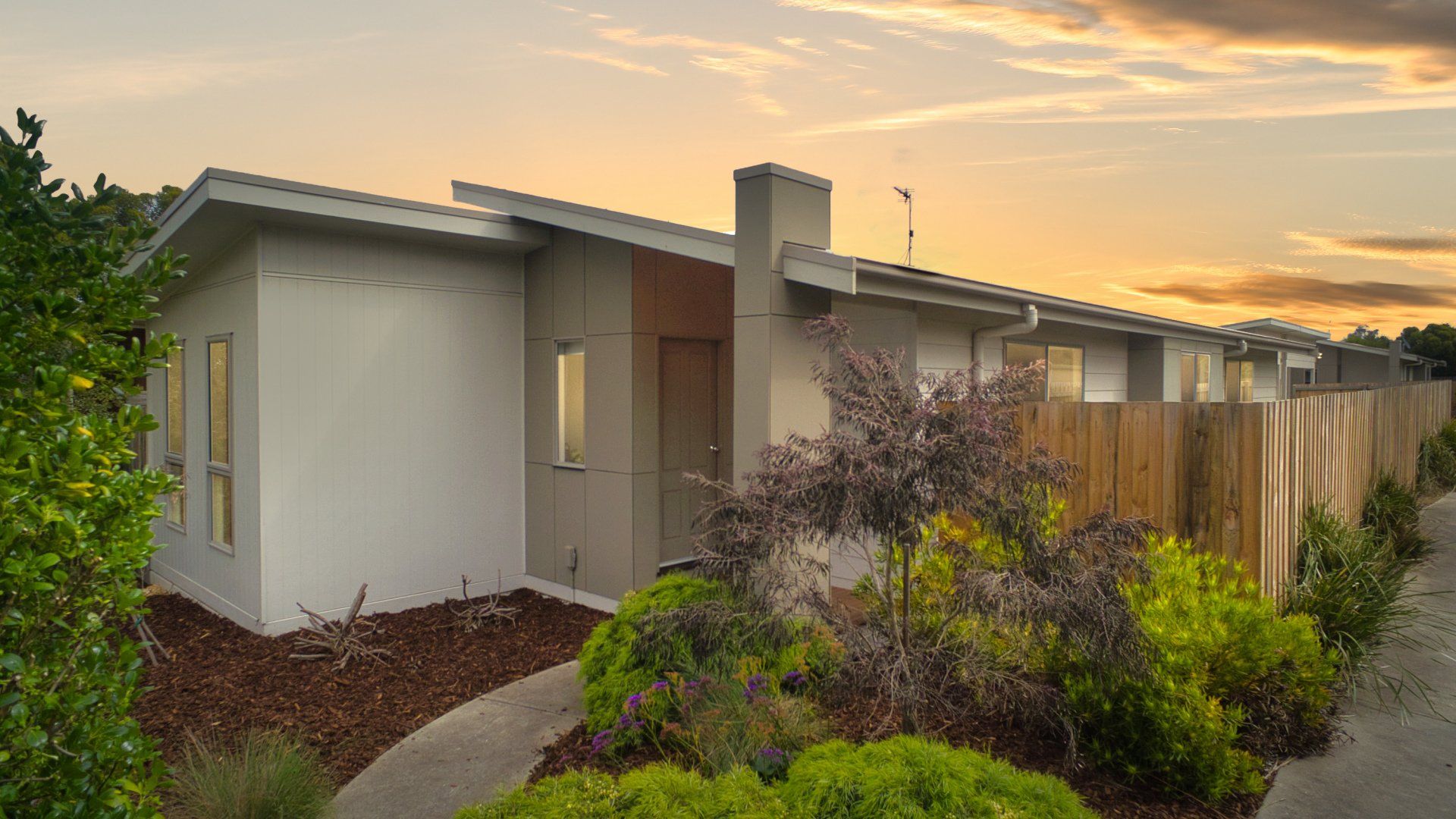 Modern single-story house with gray exterior, fenced yard, and sunset sky