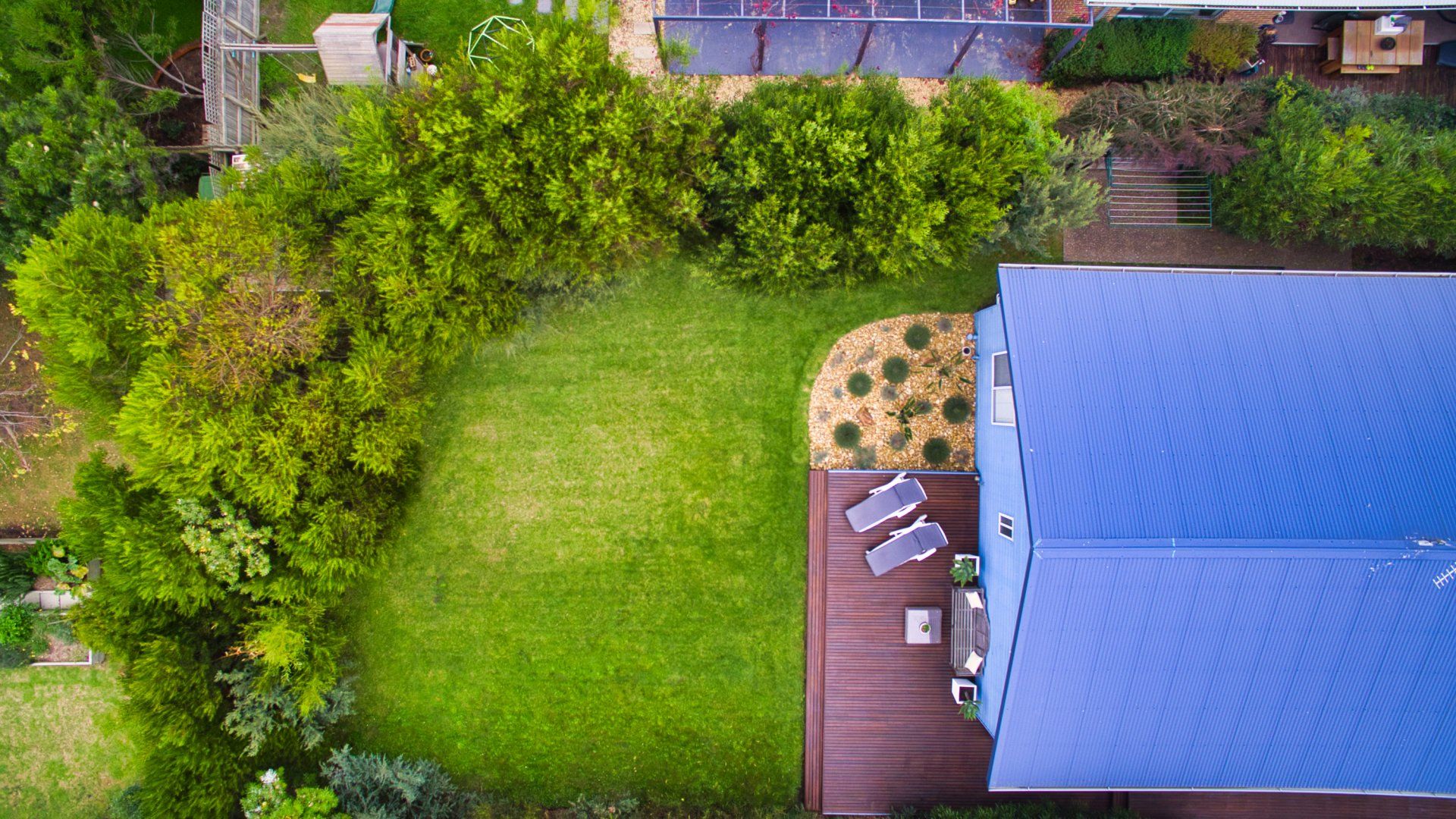 Aerial view of a backyard with green lawn, shrubs, and a blue-roofed patio beside a wooden deck.